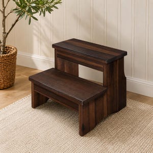 May include: A two-step wooden stool with a dark brown wood grain finish. The stool features a lower step and a higher step, both with flat rectangular surfaces, supported by solid side panels that connect the two levels into a single unit.
