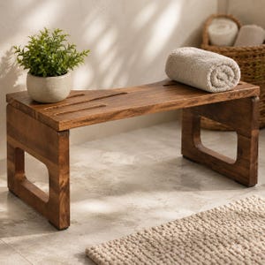 May include: A low rectangular wooden bench with a dark brown finish and a slatted top surface. A small potted green plant sits on the left side of the bench, and a rolled-up beige towel rests on the right side, featuring open-frame side supports.