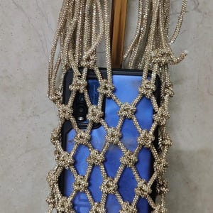 Op de afbeelding: Een blauwe smartphone in een decoratief goudkleurig nettasje met een geknoopt ruitpatroon en lange, glinsterende koordbanden. Het metalen gaas is bedekt met kleine, reflecterende kristallen die het licht opvangen.