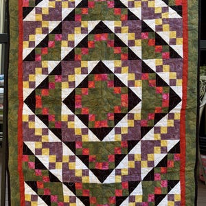 May include: A rectangular quilt featuring a geometric pattern of small squares in olive green, purple, yellow, white, and variegated red. The design includes large diamond shapes framed by a red border and an outer olive green border with black binding.