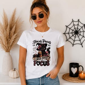 May include: A white short-sleeved t-shirt featuring a graphic of a black cat wearing a witch hat, holding a coffee cup, surrounded by books and a ghost. The text reads Hocus Pocus I NEED COFFEE TO FOCUS in black and pink leopard print lettering.