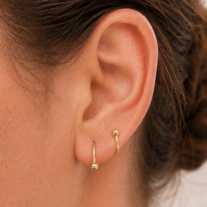 May include: A pair of gold hoop earrings, each featuring a small, polished gold sphere at the end of the wire. The earrings are worn in two separate piercings on the earlobe, with one hoop positioned slightly lower than the other, showcasing a minimalist design.