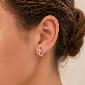 Könnte beinhalten: Ein kleiner silberfarbener Creolen-Ohrring mit einem Unendlichkeitssymbol-Design. Der Ohrring wird im Ohrläppchen getragen und zeichnet sich durch eine polierte metallische Oberfläche sowie eine durchgehende Schlaufenform aus.