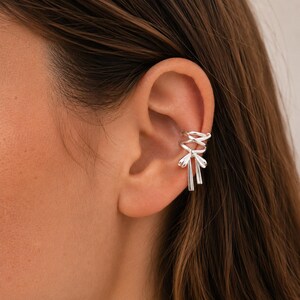 May include: A silver ear cuff featuring a polished metallic finish. The design consists of a series of interconnected loops that form a bow shape at the base, with two short, straight, dangling silver strands extending downwards from the centre knot.