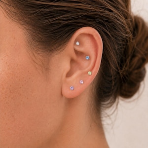 May include: A set of five small, round, prong-set stud earrings worn in a single ear. The earrings feature individual gemstones in clear, light blue, pale green, soft pink, and lavender colors, each secured with a metallic backing.
