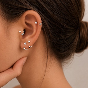 Puede incluir: Una colección de piercings de oreja en tono plateado que incluye pequeños pendientes de bola, barras curvas con extremos esféricos y un pendiente con una piedra redonda transparente. Las piezas se ubican en hélix, rook y lóbulo.