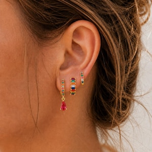 May include: Three gold-tone hoop earrings worn in a single ear. The largest hoop features a dangling pear-shaped pink stone. The middle hoop is a small band set with multi-colored rectangular stones. The smallest hoop has tiny colorful accents.