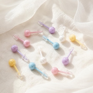 Könnte beinhalten: Eine Sammlung von Ohrsteckern mit kleinen, glatten, runden Kugeln in Pastellfarben wie Rosa, Blau, Gelb, Violett und Weiß. Jeder Ohrstecker hat einen farblich passenden Stift und einen transparenten Kunststoffverschluss für sicheren Halt.