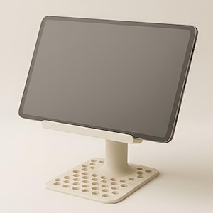 May include: A light beige tablet stand with a rectangular base featuring a grid of circular cutouts and a central support pillar. A dark grey tablet is positioned horizontally on the stand's ledge, displaying a blank, dark screen surface.