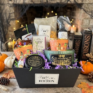 May include: A black gift box labeled The Boo Box containing a Frenchy's Collection Witches Brew candle, a Jardin Isabela tin, a Pumpkin Cream wax melt, a spray bottle, a bag of snacks, and a small jar of matches, all nestled in purple paper shred.
