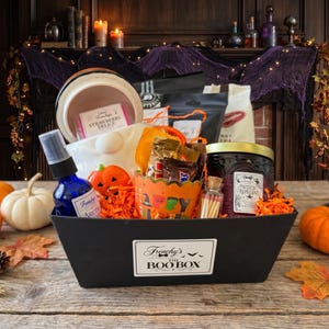 May include: A black rectangular gift basket labeled Frenchy's THE BOO BOX containing a blue spray bottle, a jar labeled Witch Repellent, a small bottle of matches, a pumpkin-shaped item, and various snacks nestled in orange paper shreds.