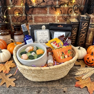 May include: A woven basket containing a light green candle in a gray concrete bowl, a blue spray bottle, a wax melt labeled A Night Affair, a black pouch, a small glass vial of matches, and an orange paper cup with Happy Halloween text.