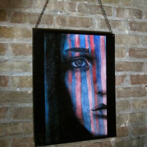 May include: A framed painting of a person's face with blue and pink stripes painted on it. The person's eye is visible and the painting is hanging on a brick wall.