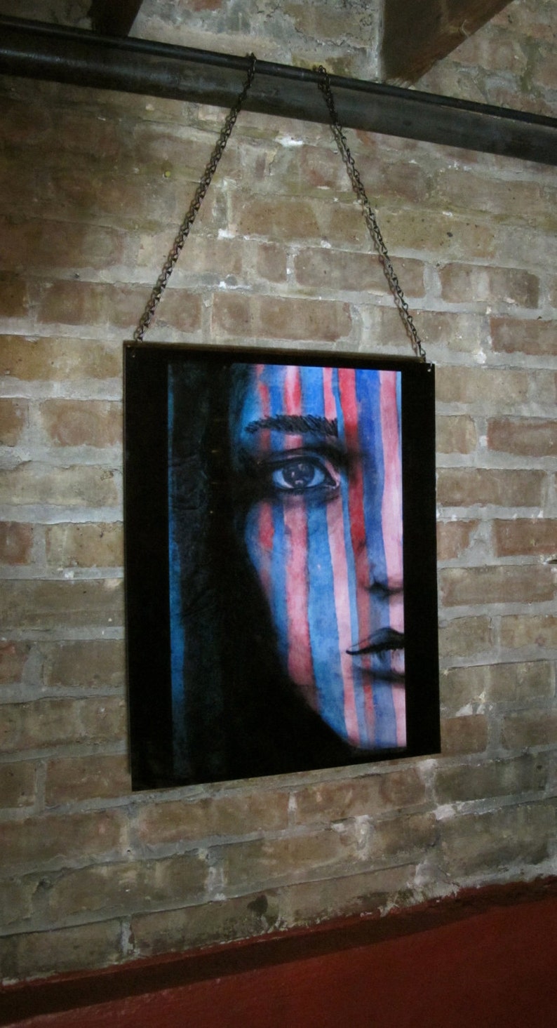 May include: A framed painting of a person's face with blue and pink stripes painted on it. The person's eye is visible and the painting is hanging on a brick wall.