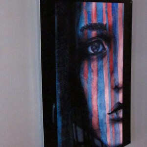 May include: A close-up of a watercolor painting of a person's face with blue and red stripes painted over the face. The person's eye is visible and the painting is framed in a black frame.