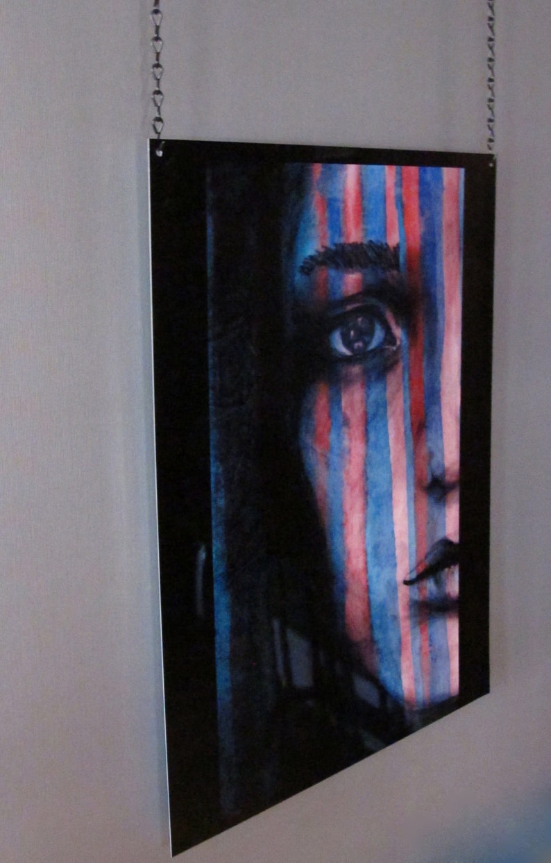 May include: A close-up of a watercolor painting of a person's face with blue and red stripes painted over the face. The person's eye is visible and the painting is framed in a black frame.