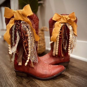 May include: A pair of red cowboy boots featuring decorative mustard yellow fabric bows tied at the top. Each boot is adorned with hanging strands of lace, ribbon, wooden beads, and textured fabric strips in shades of brown, cream, and red.