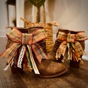 May include: A pair of brown ankle boots featuring decorative bows made from plaid fabric in shades of orange, green, and brown. The bows are accented with dangling ribbons of white lace, green fabric, and a thin strip with a small yellow floral pattern.