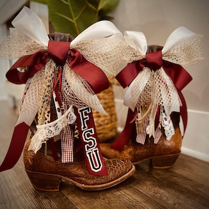May include: A pair of brown textured leather cowboy boots adorned with large decorative bows. The bows feature white satin, burgundy ribbon, and gold mesh fabric. A vertical burgundy ribbon with the white letters FSU hangs from the boots.