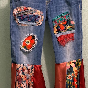 May include: Blue denim jeans featuring various fabric patches including floral, bird, and lace patterns. The lower legs are extended with panels of red satin and multi-colored floral fabric, finished with delicate lace trim at the bottom edges.