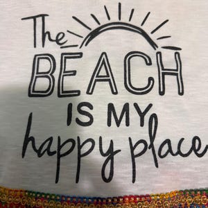 May include: A white t-shirt featuring a black graphic of a sun and the text "The BEACH IS MY happy place". The bottom hem is decorated with a colorful, multi-patterned fabric trim featuring gold, red, green, and blue woven details.