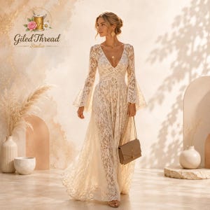 May include: A floor-length cream-colored lace dress featuring long bell sleeves, a deep V-neckline, and a fitted bodice. The dress is crafted from intricate floral lace fabric and is paired with a structured beige rectangular shoulder bag.