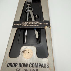 May include: A silver stainless steel Vemco drop bow compass, cat. no. D-580, housed in a gray cardboard box with a black plastic insert. The box features the text: STAINLESS STEEL, GUARANTEED WITHOUT TIME LIMIT, AMERICAN MADE, and DROP BOW COMPASS.