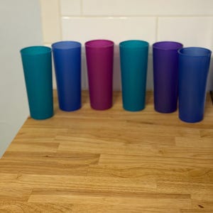 May include: A set of six tall, cylindrical drinking tumblers in a variety of translucent colors including teal, royal blue, magenta, turquoise, purple, and navy blue. Each cup features a smooth, matte finish and a simple, straight-sided design.