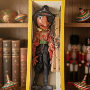 Puede incluir: Una marioneta de Pelham Puppets en una caja amarilla, con un personaje que lleva un parche negro en el ojo, una camisa roja con estampado de cachemira, una faja verde y pantalones negros. La marioneta se controla con una cruceta de madera y cuerdas.