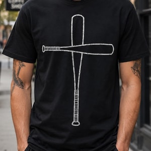 Puede incluir: Una camiseta negra de manga corta con un estampado gráfico blanco de dos bates de béisbol dispuestos en forma de cruz. El diseño tiene un aspecto desgastado y texturizado, con un bate colocado verticalmente y el otro horizontalmente.
