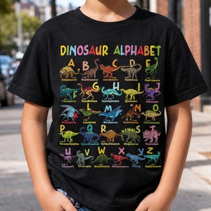 Może przedstawiać: Czarny t-shirt z kolorowym nadrukiem graficznym zatytułowanym DINOSAUR ALPHABET. Wzór przedstawia litery od A do Z, z których każda jest połączona z ilustracją dinozaura i jego nazwą, taką jak Ankylosaurus, Brachiosaurus i Zephyrosaurus.