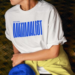 May include: A white short-sleeved t-shirt featuring the text "MORE IS MORE" in small blue letters above the large blue word "MAXIMALIST" printed in a tall, condensed, sans-serif font across the chest area of the garment.