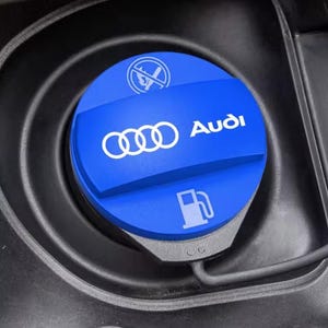 Puede incluir: Una tapa de combustible azul circular con el logotipo blanco de Audi, un icono de surtidor de gasolina y un símbolo de prohibido fumar. La tapa está situada en una carcasa negra e incluye un cable, diseñada para el depósito de un vehículo.
