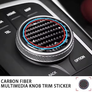 May include: A circular multimedia knob trim sticker featuring a black carbon fiber pattern with a thin red inner ring and a light blue outer ring. The sticker has a textured surface and is designed to fit over a car's central control dial.
