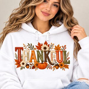 Peut inclure: Un sweat-shirt à capuche blanc avec le mot THANKFUL imprimé en grandes lettres majuscules multicolores. Le motif est décoré d'illustrations sur le thème de l'automne, notamment des citrouilles orange, des glands marron, des tournesols et des feuilles.