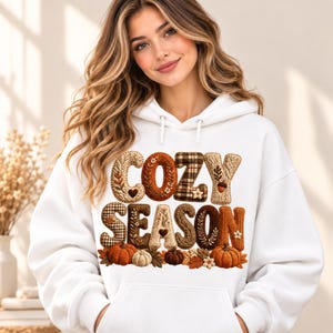 May include: A white hooded sweatshirt featuring the text COZY SEASON in large, textured letters with various autumn-inspired patterns including plaid, cable knit, and floral motifs, accented by small pumpkins and fall leaves at the base.