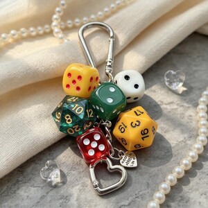 May include: A keyring featuring a silver-toned metal carabiner and a heart-shaped clasp. The charm assembly includes a yellow die, a white die, a green die, a red die, a translucent green twenty-sided die, and a yellow twenty-sided die.