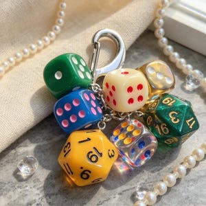 Puede incluir: Un llavero con un cierre de metal plateado unido a un grupo de dados de varios colores. La colección incluye dados de seis caras en verde, azul, amarillo y crema, un dado translúcido multicolor y dos dados de veinte caras numerados.