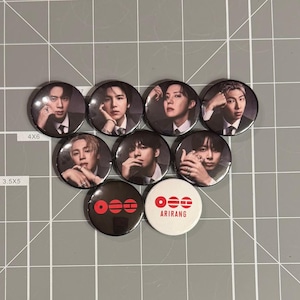 May include: A collection of nine circular pin-back buttons featuring portraits of seven individuals in dark suits, alongside two additional buttons. One button is black with red geometric symbols, and one is white with red symbols and the text ARIRANG.