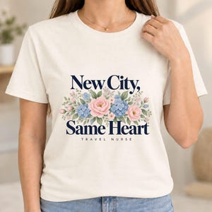 Może przedstawiać: Kremowy t-shirt z krótkim rękawem z ciemnoniebieskim napisem New City, Same Heart oraz TRAVEL NURSE. Wzór zawiera centralną kompozycję kwiatową z różowymi różami, niebieskimi kwiatami przypominającymi hortensje i zielonymi liśćmi.