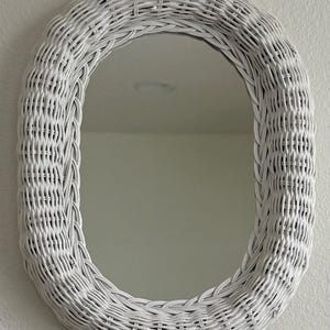 May include: An oval-shaped wall mirror featuring a thick, woven white wicker frame. The mirror glass is clear and reflective, set within the center of the textured, braided white wicker border that surrounds the entire perimeter of the piece.