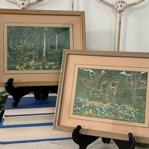 May include: Two rectangular framed prints featuring detailed landscape illustrations of lush green forests, meadows with colorful wildflowers, and wooden fences, each encased in a light tan frame with a matching matting border.