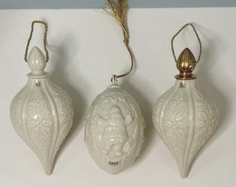 May include: Three off-white ceramic ornaments with gold hanging cords. Two are teardrop-shaped with embossed floral patterns, one with a gold cap. The center ornament is oval-shaped with a raised Santa Claus figure and the year 1995 printed on it.