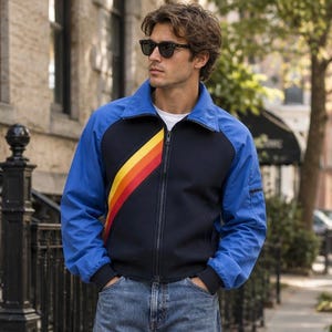 Puede incluir: Una chaqueta con cremallera que presenta un torso azul marino con un patrón de rayas diagonales en amarillo, naranja y rojo, complementada con mangas y cuello en azul brillante, con cremallera frontal, sobre una camisa blanca y vaqueros.