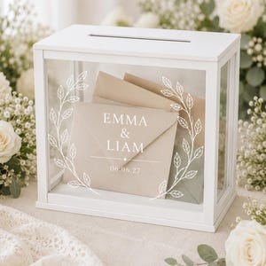 Op de afbeelding: Een rechthoekige kaartenbox van wit hout met doorzichtige glazen panelen. Het voorste glas is voorzien van een wit gedrukt botanisch kransontwerp rondom de tekst EMMA & LIAM en de datum 06.06.27. Binnenin zijn verschillende beige enveloppen zichtbaar.