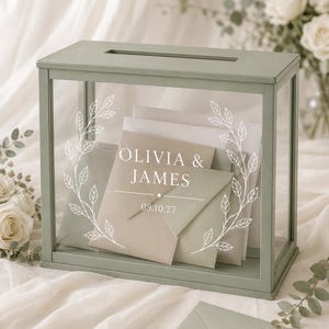 Könnte beinhalten: Eine rechteckige Kartenbox mit einem salbeigrünen Holzrahmen und klaren Glasscheiben. Auf der vorderen Glasscheibe befindet sich ein weißer Lorbeerkranz-Aufdruck, die Namen OLIVIA & JAMES sowie das Datum 05.10.27. Im Inneren befinden sich neutrale Umschläge.