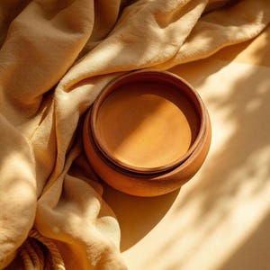 May include: A round, terracotta-colored clay bowl with a smooth, matte finish and a wide, open top. The bowl features a simple, circular design with a consistent earthy tone, resting near a draped, soft, light-tan fabric with visible folds.