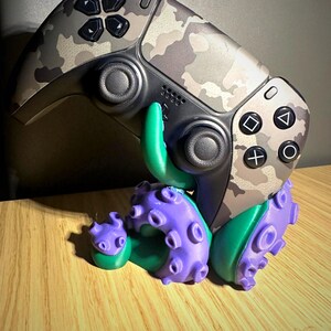 Puede incluir: Un mando de videojuegos con estampado de camuflaje sostenido por un soporte con forma de pulpo morado y verde. El mando cuenta con joysticks negros, una cruceta negra y botones de acción estándar. El soporte tiene ventosas texturizadas.