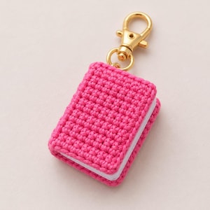 Può includere: Un portachiavi a forma di libro in miniatura con una copertina all'uncinetto rosa acceso dal motivo testurizzato. Il libro presenta pagine bianche visibili sul lato ed è fissato a un moschettone in metallo color oro lucido.