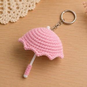 May include: A pink crocheted umbrella keyring featuring a scalloped edge canopy, a white central shaft, and a matching pink crocheted handle. The umbrella is attached to a silver-toned metal split ring by a short chain, creating a small accessory.